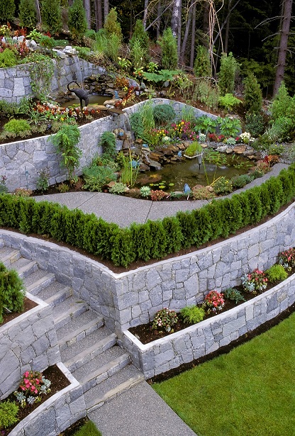 Tiered stone retaining walls with garden beds, a pond and steps in a landscaped backyard