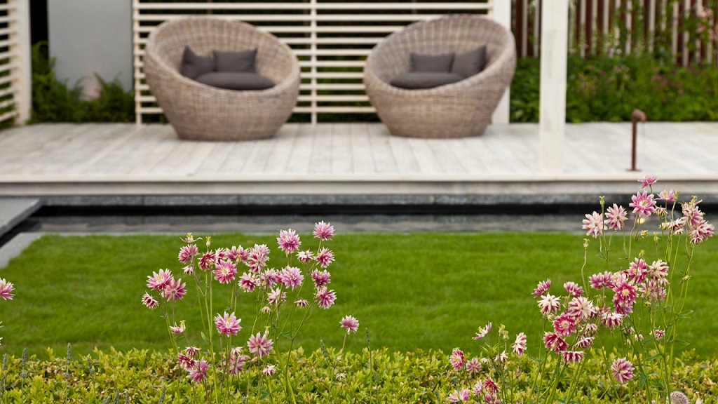 Wicker outdoor chairs on a deck beside a lawn with pink flowering plants